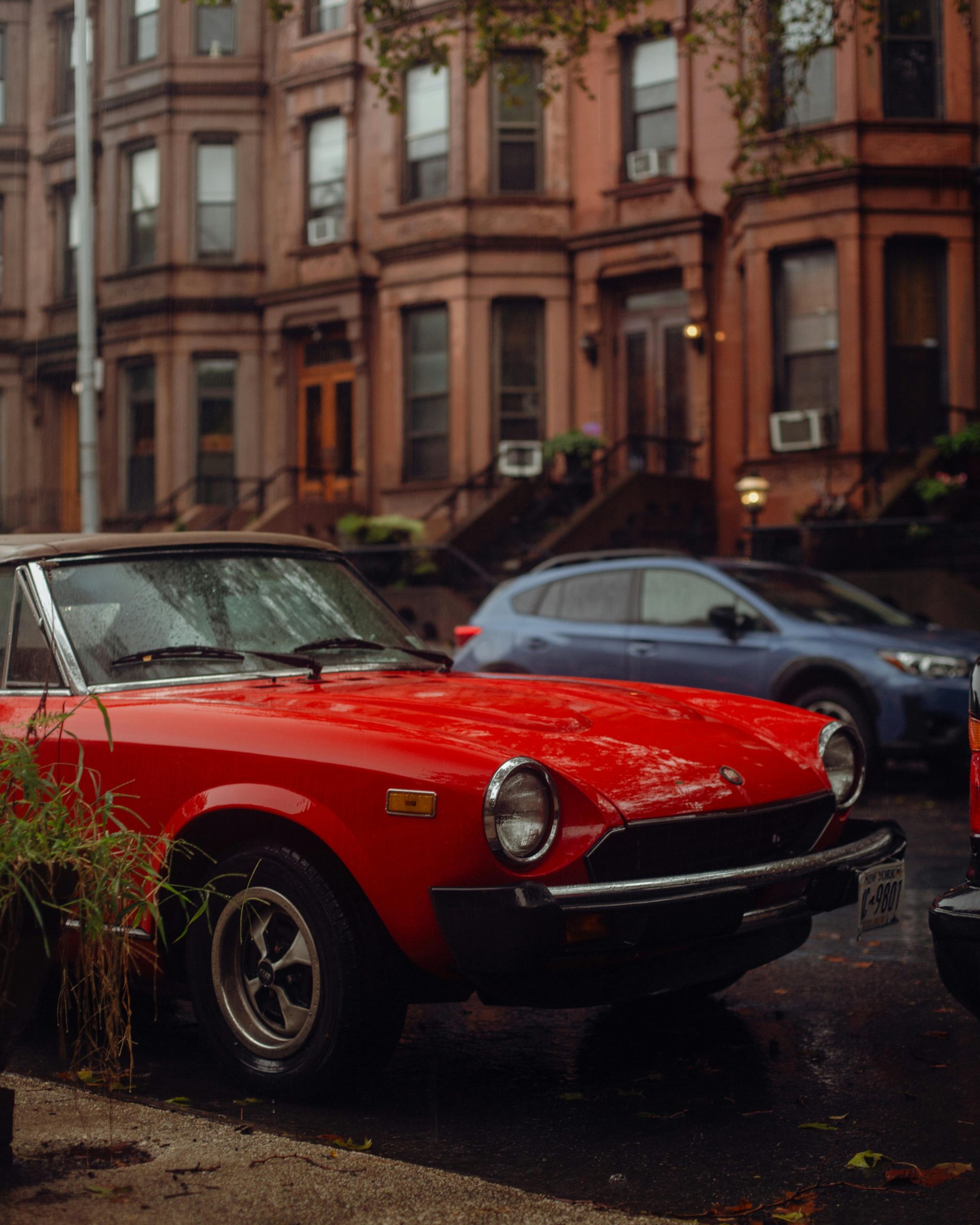 Classic red Fiat Spider garée dans une rue urbaine pluvieuse avec des bâtiments historiques.