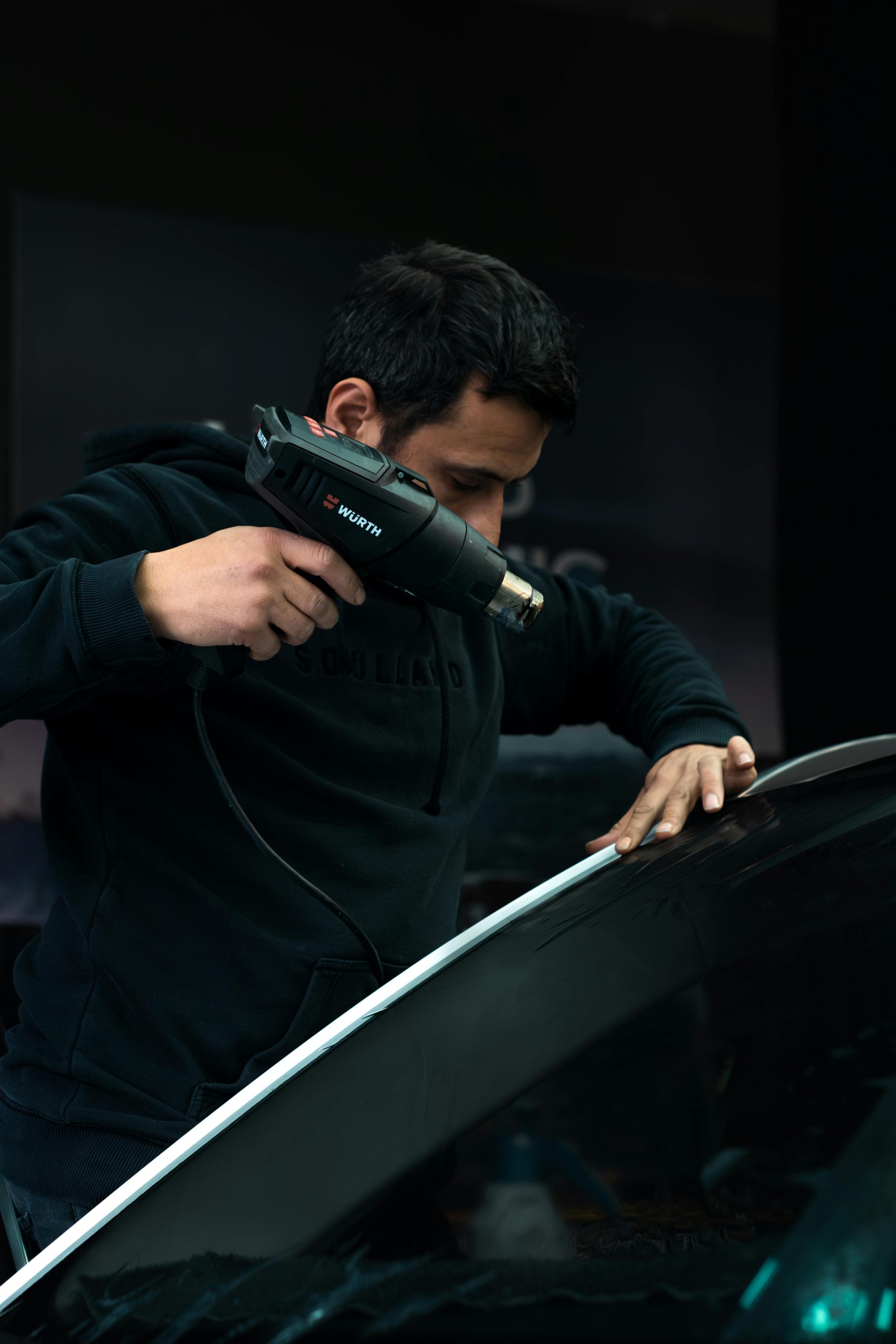 Mécanicien appliquant soigneusement de la chaleur sur un pare-brise de voiture avec un pistolet thermique dans un garage.