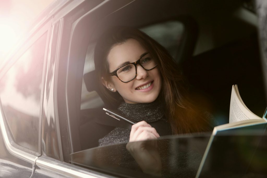 Smiling young woman reading a book in a car, enjoying leisure time during a sunny day. Une jeune femme souriante lisant un livre dans une voiture, profitant du temps libre par une journée ensoleillée.