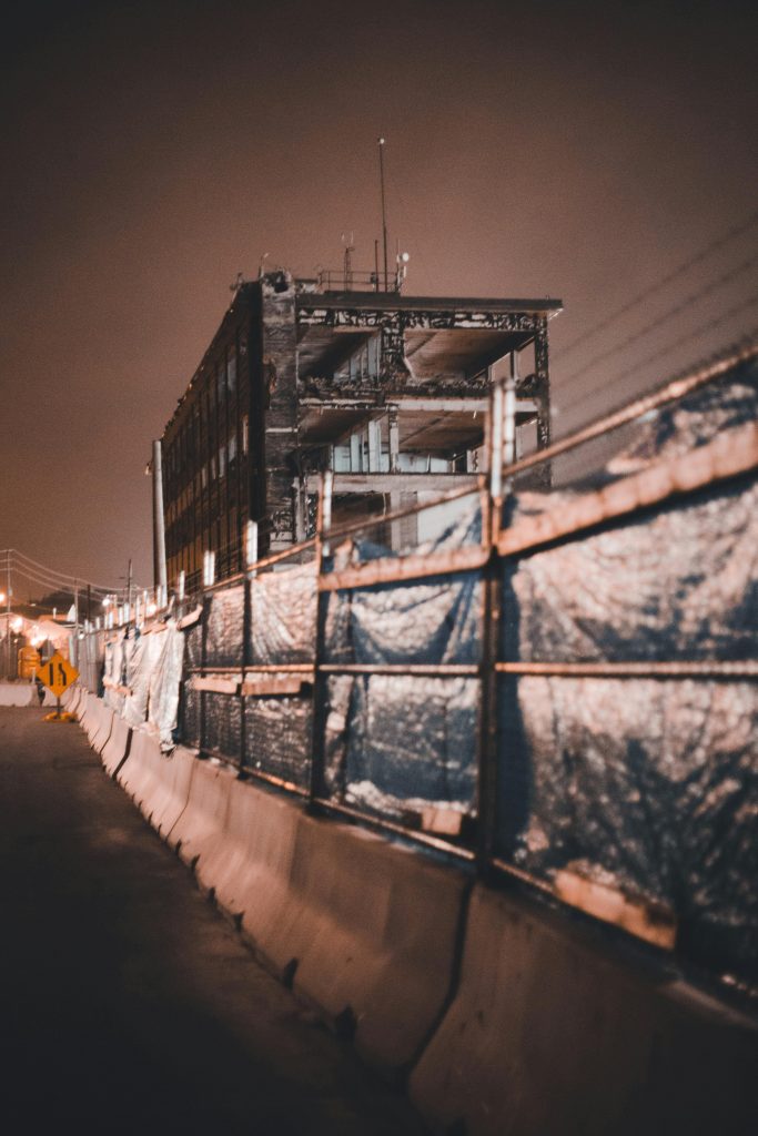 A broken building with concrete barriers and metal fences under a dark, night sky. Un bâtiment détruit avec des barrières en béton et des clôtures métalliques sous un ciel nocturne sombre.