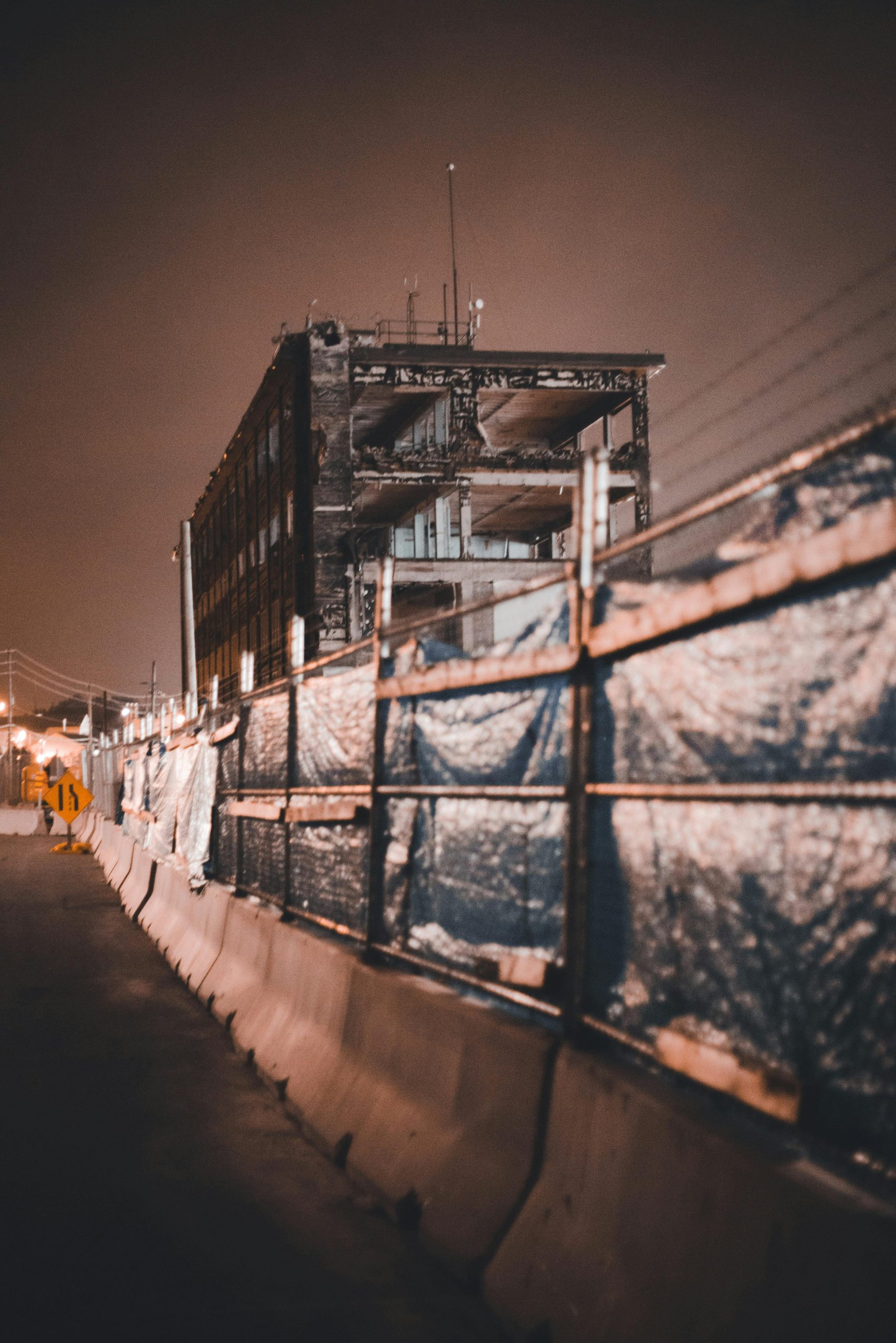 A broken building with concrete barriers and metal fences under a dark, night sky. Un bâtiment détruit avec des barrières en béton et des clôtures métalliques sous un ciel nocturne sombre.