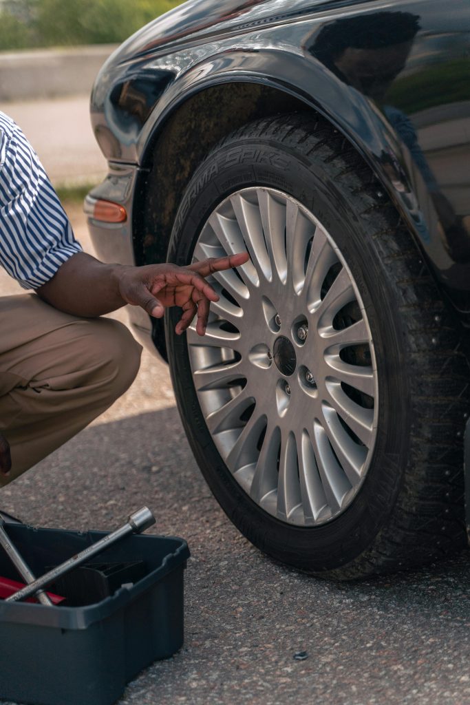 An adult male pointing at a car tire, suggesting a focus on vehicle maintenance or inspection outdoors. Un homme adulte pointant du doigt un pneu de voiture, suggérant une attention particulière à l'entretien ou à l'inspection des véhicules en extérieur.