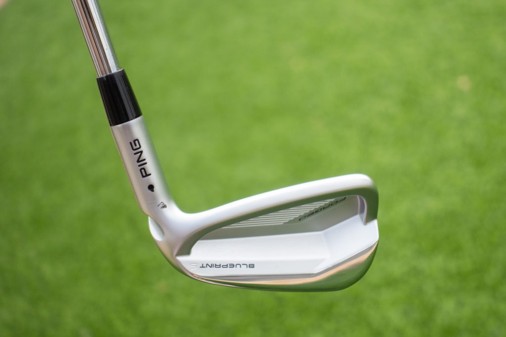 High-quality image of a Ping Blueprint golf iron on a grassy background. Image de haute qualité d'un fer de golf Ping Blueprint sur un fond herbeux.