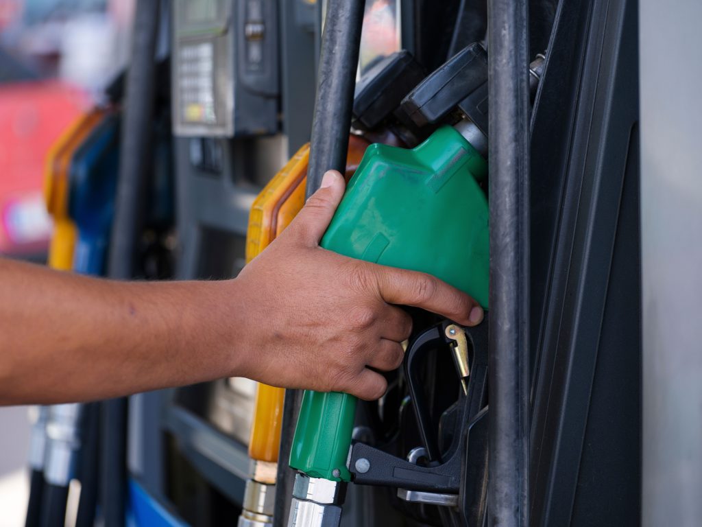 Close-up of a hand holding a green fuel nozzle at a gas pump station outdoors. Gros plan d'une main tenant un pistolet de carburant vert à une pompe à essence en extérieur.