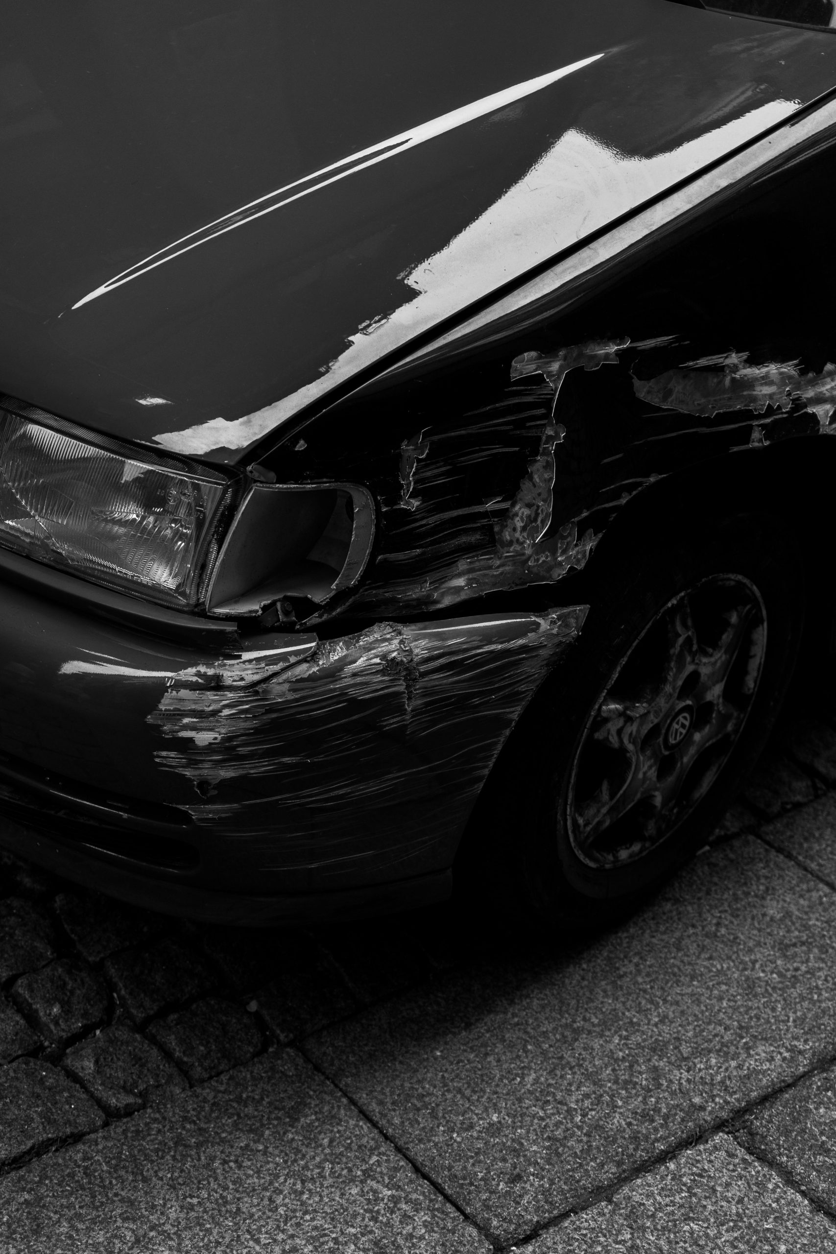 Artistic black and white image of a scratched car fender parked on a cobblestone sidewalk in Porto. Image artistique en noir et blanc d'une aile de voiture rayée garée sur un trottoir en pavés à Porto.
